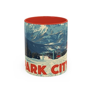 Może przedstawiać: Biały ceramiczny kubek z czerwonym wnętrzem. Ilustracja ośnieżonego pasma górskiego i wyciągów narciarskich. Napis "PARK CITY" w czerwonych literach.
