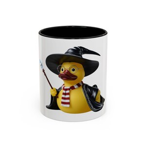 May include: A white ceramic mug with a black rim features a cartoon image of a yellow rubber duck wearing a black wizard hat and glasses, holding a wand. The duck is wearing a red and white striped scarf and a black jacket.