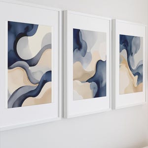 Puede incluir: Tres impresiones artísticas abstractas enmarcadas. Cada impresión presenta formas fluidas en tonos azul marino, azul claro y beige. La obra de arte está enmarcada en blanco, creando una estética moderna y minimalista. Las impresiones se exhiben en una pared blanca.