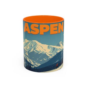 May include: A ceramic coffee mug with an orange interior and the word "ASPEN" in orange block letters. The mug features a scenic illustration of snow-capped mountains, a skier, and a blue sky.