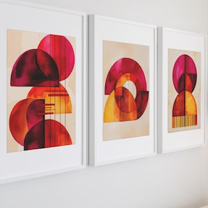 Puede incluir: Tres impresiones artísticas abstractas enmarcadas. Cada impresión presenta formas geométricas superpuestas en tonos rojos, naranjas y amarillos sobre un fondo neutro. Los marcos son blancos. La obra está en una pared blanca.