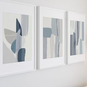 Puede incluir: Tres impresiones artísticas abstractas enmarcadas. Cada impresión presenta diseños de estilo pincelada en tonos de azul, gris y blanco. Los marcos son blancos y la obra de arte se exhibe en una pared blanca.