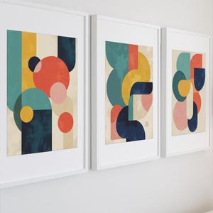 Puede incluir: Tres impresiones artísticas abstractas enmarcadas. Cada impresión presenta un diseño geométrico único con formas en verde azulado, amarillo, azul marino, coral y rosa. Las impresiones se muestran en marcos blancos contra una pared blanca.