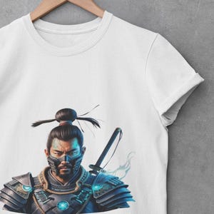 Può includere: Maglietta bianca con una stampa grafica di un guerriero samurai che indossa una maschera e un'armatura blu e nera, tenendo una spada.