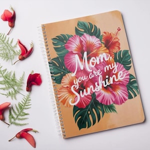 Puede incluir: Una libreta de espiral con un diseño floral con flores de hibisco rosas y rojas y hojas verdes. La libreta tiene el texto "Mom, you are my sunshine" impreso en la portada.