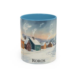 Puede incluir: Una taza de café de cerámica con interior azul claro y una escena de paisaje invernal. La escena representa casas coloridas en un pueblo nevado con la palabra "ROROS" impresa en la parte inferior.