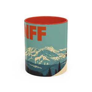 May include: A ceramic coffee mug with a red interior and a teal exterior featuring the word "BANFF" in orange block letters. The design includes a mountain range, a ski lift, and pine trees.