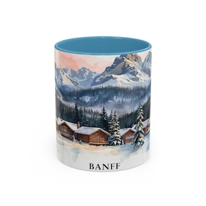 May include: A ceramic coffee mug with a blue interior and a wraparound watercolor design. The design features a mountain range, evergreen trees, and cabins in a snowy landscape. The word "BANFF" is printed on the bottom.