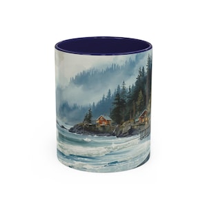 May include: A white ceramic mug with a blue interior features a watercolor painting of a coastal scene. The artwork depicts a rocky shoreline with cabins, evergreen trees, and a misty mountain backdrop. The waves crash against the shore.