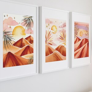 Puede incluir: Tres pinturas de acuarela enmarcadas que representan un paisaje desértico con colinas onduladas, una puesta de sol y palmeras. Las pinturas están en tonos de rosa, naranja y marrón.