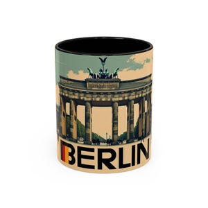 May include: A ceramic coffee mug with a black interior and a design featuring the Brandenburg Gate in Berlin. The mug has the word "BERLIN" in large letters with the German flag colors.