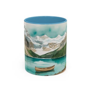 May include: A ceramic coffee mug featuring a watercolor painting of a mountain lake scene. The design includes snow-capped mountains, a turquoise lake, a small wooden boat, and green trees. The interior of the mug is light blue.