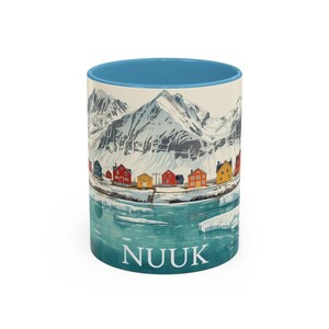 May include: A ceramic mug with a blue interior and a scenic illustration. The design features a snowy mountain range, colorful houses, and the word "NUUK" in white. The mug is a standard size.