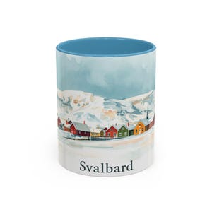 Puede incluir: Taza de cerámica blanca con interior azul y una ilustración de acuarela de un paisaje nevado con casas coloridas. La palabra "Svalbard" está impresa en la parte inferior de la taza.