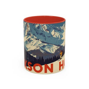 May include: A white ceramic mug with a red interior featuring a vintage-style illustration of a snowy mountain range and a skier. The text "JACKSON HOLE" is printed in red.