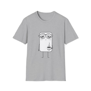 Puede incluir: Camiseta gris con un personaje de dibujos animados. El personaje es un rectángulo blanco con contornos negros, ojos somnolientos y una taza de café. El diseño está centrado en la parte delantera de la camiseta, con el personaje sosteniendo una taza de café humeante.