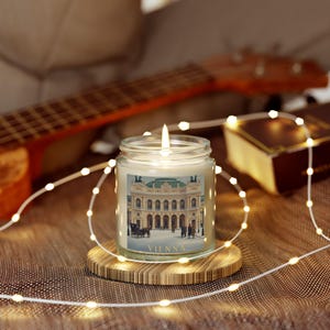 Puede incluir: Una vela encendida en un tarro de cristal con una etiqueta que presenta una ilustración vintage de un edificio con la palabra "VIENNA" en la parte inferior. La vela está sobre un posavasos de madera, rodeada de luces de cadena y un ukelele.