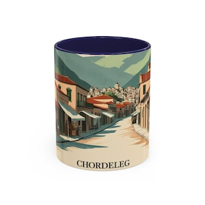 May include: A ceramic mug with a blue interior, featuring a wraparound illustration of a street scene in Chordeleg, Ecuador. The design shows buildings with red tile roofs, a cobblestone street, and mountains. The word "CHORDELEG" is printed on the mug.