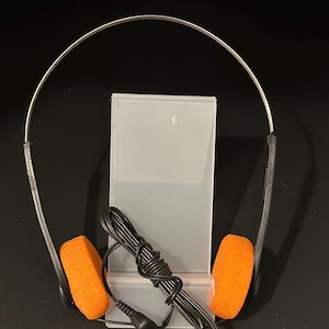 Puede incluir: Auriculares plateados con almohadillas naranjas brillantes y un cable negro. Los auriculares están sobre un soporte de plástico transparente sobre un fondo oscuro. Las almohadillas son rectangulares y de material poroso.