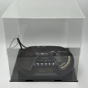 Puede incluir: Un radio reloj Sony Dream Machine negro dentro de una vitrina de acrílico transparente. La radio tiene una pantalla digital y varios botones. La vitrina se encuentra sobre una base negra.