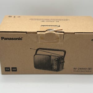 Könnte beinhalten: Brauner Karton mit dem Panasonic-Logo und der Modellnummer RF-2400D. Die Box zeigt eine Grafik eines tragbaren Radios mit Antenne und den FM/AM-Radiobändern.