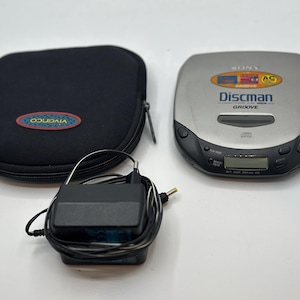 Può includere: Un lettore CD Sony Discman argentato con dettagli neri, una custodia nera e un adattatore di alimentazione nero. Sul Discman è stampata la scritta "Discman". Sulla custodia è stampata la scritta "Vivanco".