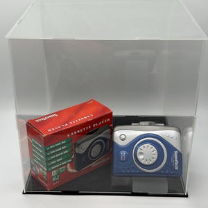 Puede incluir: Una caja roja y blanca con la etiqueta "Cassette Player" y un reproductor de casetes azul y plateado se exhiben dentro de una caja de acrílico transparente. El reproductor de casetes tiene la marca "Supertech" impresa.