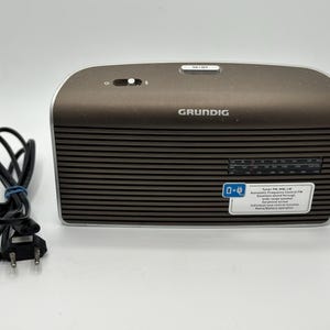 Könnte beinhalten: Ein Grundig-Radio mit braunem Oberteil und einer schwarzen, horizontal gestreiften Vorderseite. Das Radio hat ein Einstellrad und einen kleinen Aufkleber mit Text. Ein schwarzes Netzkabel ist links aufgerollt.