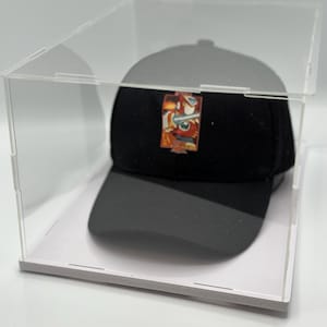 May include: A black baseball cap with a colourful graphic design is displayed in a clear acrylic display case with a white base.