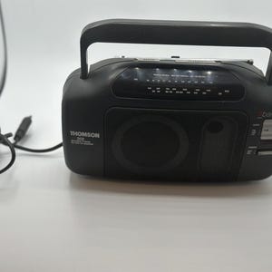 Peut inclure: Radio portable Thomson 302 noire avec une poignée, un haut-parleur et une antenne. La radio a un cadran de réglage et un cordon d'alimentation. La radio est sur une surface blanche.