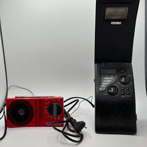 May include: Two radios on a white surface. A small red AM radio with a black dial and speaker is next to a larger black radio with a flip-up light. The red radio has the word "INTERNATIONAL" printed on it.