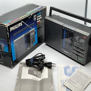 Puede incluir: Una radio portátil Philips D2225 de 5 bandas vintage, gris oscuro con asa negra, junto con su caja y manual originales. La caja es negra y azul, con el logotipo de Philips y las especificaciones de la radio. La radio tiene un mapa del mundo.