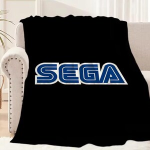 Op de afbeelding: Zwarte deken met het SEGA-logo in wit en blauw. De deken is over een lichtgekleurde bank gedrapeerd. Het SEGA-logo is een gestileerd woordmerk, een klassiek ontwerp geassocieerd met videogames.