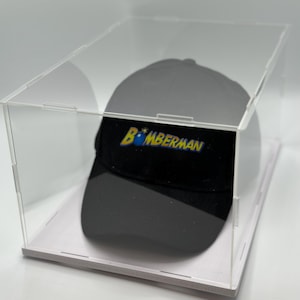 May include: A black baseball cap with a white and yellow embroidered logo that says "Bomberman" with a cartoon bomb graphic. The cap is displayed in a clear acrylic display case with a white base.