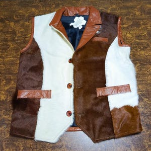 Può includere: Un gilet patchwork con sezioni di pelliccia marrone, bianca e marrone scuro. Il gilet ha un colletto in pelle marrone, dettagli delle tasche e chiusure con bottoni. La fodera interna è in tessuto blu scuro.