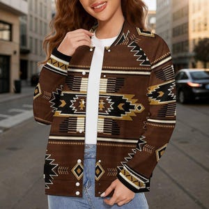 May include: A brown bomber jacket with a geometric pattern in black, white, and gold. The jacket has a button-up front and a ribbed collar. The design features diamond and zigzag shapes. The jacket is worn over a white shirt and blue jeans.