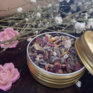 May include: A round gold-colored metal tin filled with a mixture of dried flower petals, lavender buds, and herbs. Beside the open tin lies its gold lid, which features a circular label with text. Two small pink wax roses are placed nearby.