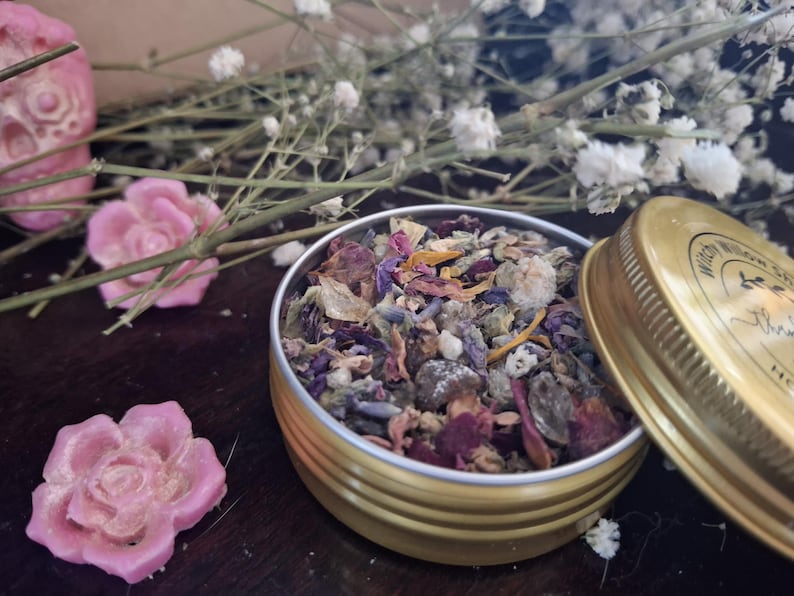 May include: A round gold-colored metal tin filled with a mixture of dried flower petals, lavender buds, and herbs. Beside the open tin lies its gold lid, which features a circular label with text. Two small pink wax roses are placed nearby.