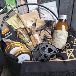 May include: A black metal cauldron filled with various items including a bottle labelled Fountain of Youth Water, two gold-lidded jars, a small broom, a glass vial with dried herbs, a black cloth with gold pentagrams, and several small boxes and pouches.