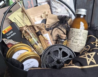 May include: A black metal cauldron filled with various items including a bottle labelled Fountain of Youth Water, two gold-lidded jars, a small broom, a glass vial with dried herbs, a black cloth with gold pentagrams, and several small boxes and pouches.