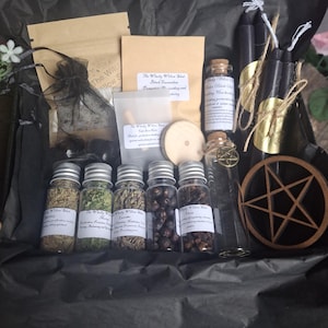 May include: A gift box containing five small glass jars filled with dried herbs and berries, two black taper candles with gold labels, a wooden pentacle, a small glass vial with a metal charm, and several paper pouches with printed labels.