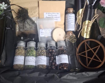 May include: A gift box containing five small glass jars filled with dried herbs and berries, two black taper candles with gold labels, a wooden pentacle, a small glass vial with a metal charm, and several paper pouches with printed labels.