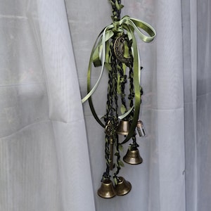 May include: A decorative hanging ornament featuring braided black and green cord intertwined with green ribbon. It includes brass-colored bells, a metal crescent moon charm, a small skeleton key, and a tiny glass vial filled with small particles.