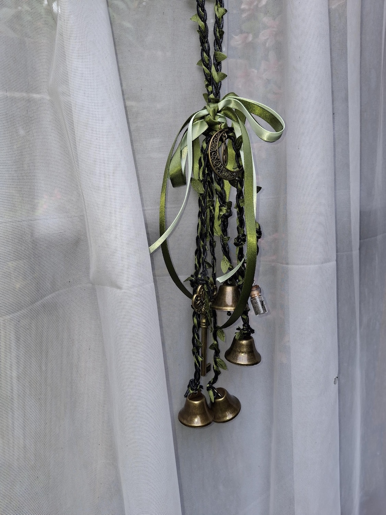 May include: A decorative hanging ornament featuring braided black and green cord intertwined with green ribbon. It includes brass-colored bells, a metal crescent moon charm, a small skeleton key, and a tiny glass vial filled with small particles.