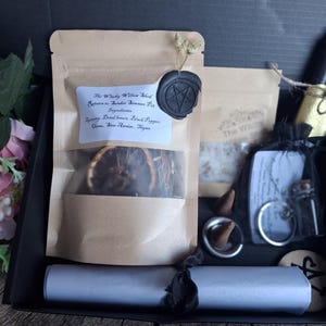 May include: A black gift box containing a tan pouch with a black wax seal, a smaller tan pouch, two black candles with gold bases, a black organza bag with a small glass vial and keychain, two incense cones, and a rolled silver paper scroll.