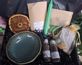 May include: A gift box containing a dried orange slice, a green taper candle, two small glass vials with dried herbs, a pouch of green crystals, a gold organza bag with two polished stones, and a small, round, speckled blue-green ceramic bowl.