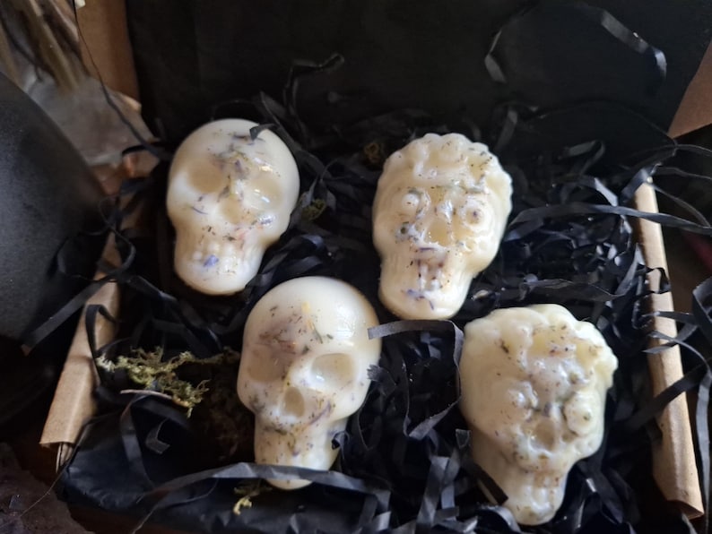May include: Four off-white skull-shaped wax melts embedded with dried botanical pieces. The wax skulls are arranged inside a cardboard box filled with black shredded paper filler, showcasing detailed facial features and textured surfaces.