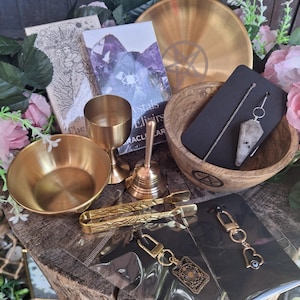 May include: A collection of ritual items including a gold-toned bowl, a chalice, a bell, and a wooden bowl containing a crystal pendulum on a chain. Also included are gold-toned tongs, two keychains with charms, and a deck of oracle cards.