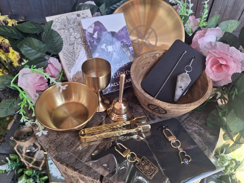 May include: A collection of ritual items including a gold-toned bowl, a chalice, a bell, and a wooden bowl containing a crystal pendulum on a chain. Also included are gold-toned tongs, two keychains with charms, and a deck of oracle cards.