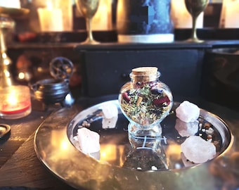 May include: A heart-shaped glass bottle with a cork stopper, filled with a mixture of dried herbs, flower petals, and small dark berries. The bottle sits on a round silver-toned metal tray surrounded by four rough-cut, pale pink quartz stones.